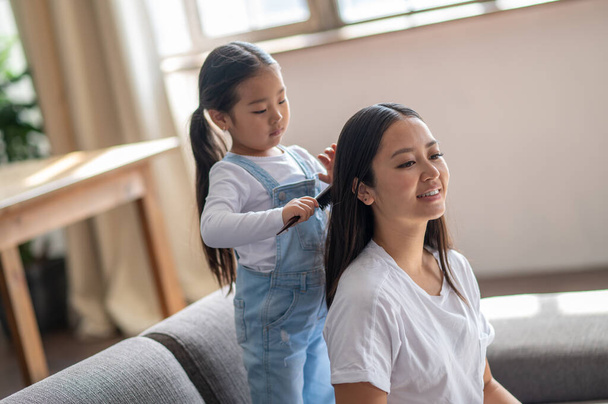 Serious little Asian girl standing on the sofa and brushing her pleased female parent hair - Foto, immagini