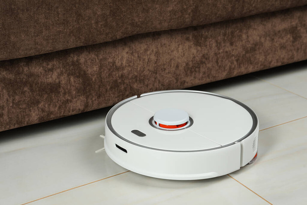 Robotic vacuum cleaner on white tiled floor near sofa indoors - Photo, image