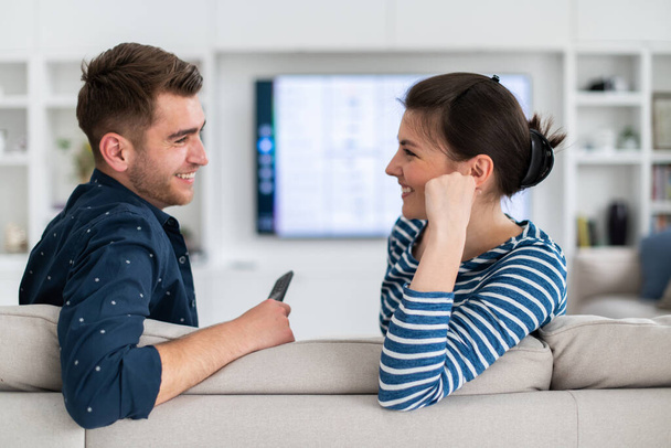 Couple Watches TV together while Sitting on a Couch in the Living Room. Girlfriend and Boyfriend embrace, cuddle, talk, smile and watch Television Streaming Services. Home with Cozy Stylish Interior - Фото, изображение