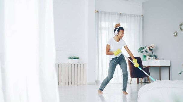 Pretty African American girl cheerful maid is cleaning floor in beautiful flat with plastic mop and listening to music, singing and dancing. Routine, house and fun concept. - 写真・画像