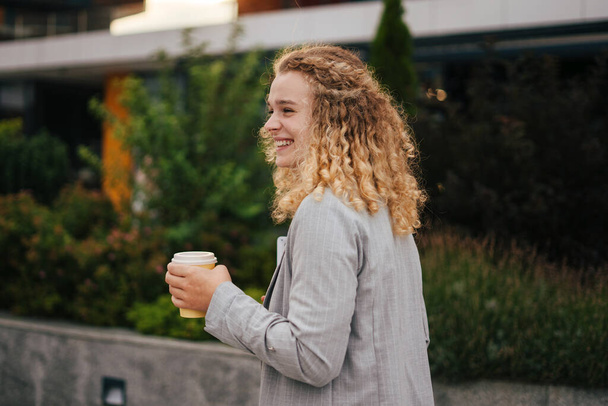 Sonriendo joven estudiante, caminando por la calle mientras bebe café en una taza y sosteniendo un portátil en sus manos. Estilo de vida moderno. Gente moderna y exitosa - Foto, Imagen