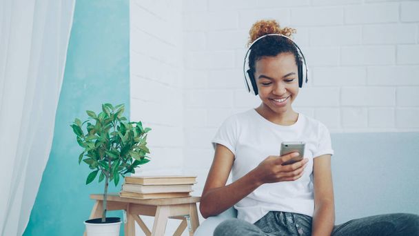 Happy African American woman is surfing internet with smartphone and laughing sitting on comfortable sofa at home, girl is listening to music through headphones. - Foto, Imagem