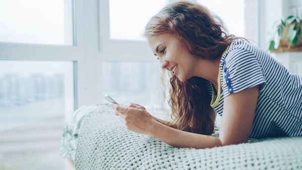 Attractive young lady is using smartphone lying on bed at home, touching screen and smiling. Beautiful light apartment with panoramic windows and wooden furniture is visible. - 写真・画像