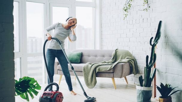 Beautiful young woman is hoovering the floor at home using modern vacuum cleaner and listening to music with headphones, dancing and singing. Housework and technology concept. - Photo, image