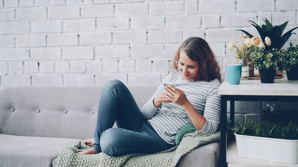Atraktivní dívka v neformálním oblečení používá smartphone sedět na pohodlné pohovce v moderní Loftový styl byt a usmívá se. Moderní technologie, koncept interiéru a mládež. - Fotografie, Obrázek