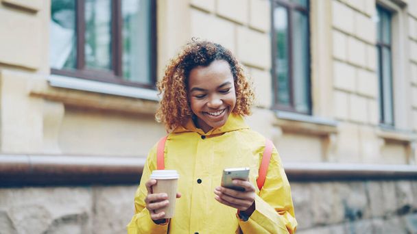 Attractive young African American woman is using modern smartphone looking at screen and smiling surfing the net or watching photos and holding take away coffee standing outdoors. - Фото, зображення