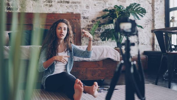Emotional young woman vlogger is talking and gesturing recording video with professional camera for internet blog. Girl is wearing casual clothing and sitting on bedroom floor at home. - Photo, Image