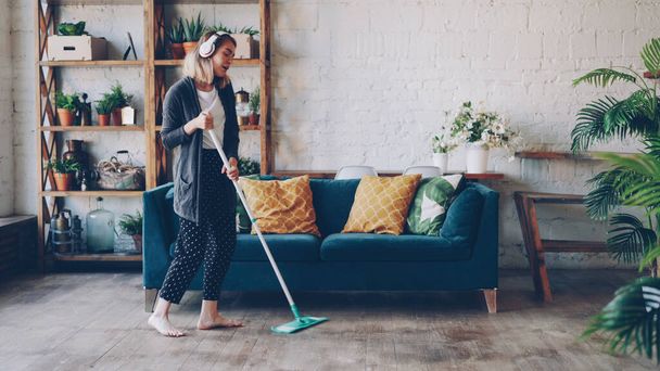Cheerful girl is mopping floor and listening to music with wireless headphones, she is singing and dancing enjoying rhythm cleaning her nice modern apartment. Joy and routine concept. - Фото, изображение