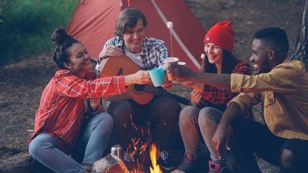 Joyful young people friends are clinking glasses with drinks sitting around fire nera tent in forest with warm marshmallow on sticks, smiling man is holding guitar. - Foto, Imagem