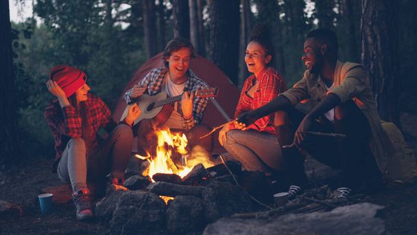 cheerful guy in casual clothing is playing the guitar while his male and female friends tourists are singing and smiling sitting around fire in forest in summer. - Fotó, kép