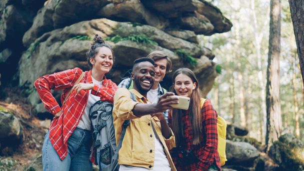 Funny young people happy friends are taking selfie in wood with mossy rocks in background, African American guy is holding smartphone, smiling men and women are posing. - Photo, image