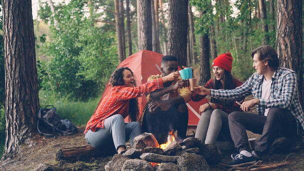 Carefree tourists are toasting, clinking cups and glasses with hot drinks then drinking sitting around fire in forest near tent. Friendship, recreation and youth concept. - Foto, Imagem
