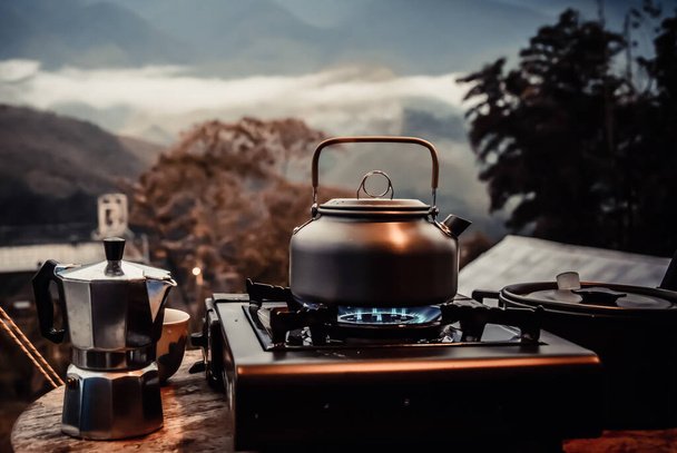 Kültéri konyha berendezések tábor tűz és főzés teáskanna moka kávé csepegtető csésze, fa asztal kemping gáz tűzhely a természetben szabadban, hegyi háttér, koncepció tábor és utazás, vintage árnyalatú - Fotó, kép