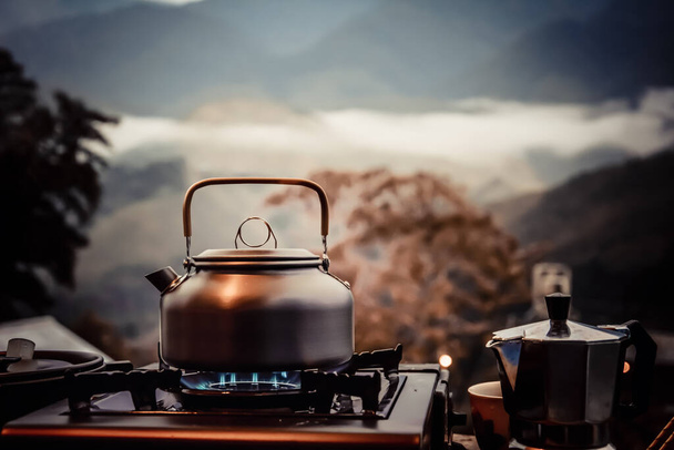 Venkovní kuchyňské vybavení tábor oheň a vaření čaj hrnec moka káva kapací šálek, s dřevěným stolem kempu plynový sporák v přírodě venkovní, horské zázemí, koncept tábor a cestování, vintage tón barva - Fotografie, Obrázek