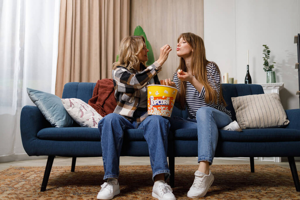 volwassen moeder en haar volwassen dochter voeden elkaar met popcorn zitten op de bank in gezellig appartement - Foto, afbeelding