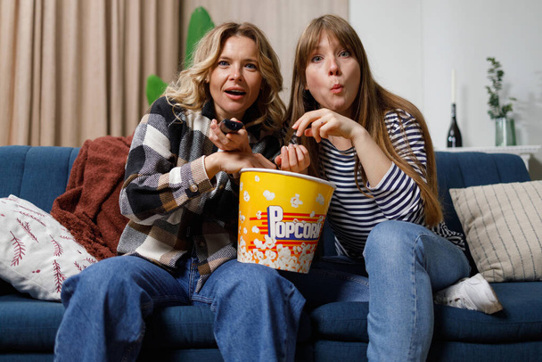 Mother and daughter are concentrating on watching an interesting movie and eating popcorn while sitting on the couch - Foto, immagini