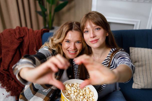 Dwie szczęśliwe kobiety pokazują znak serca dłońmi siedząc na kanapie z popcornem - Zdjęcie, obraz