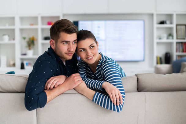 Couple Watches TV together while Sitting on a Couch in the Living Room. Girlfriend and Boyfriend embrace, cuddle, talk, smile and watch Television Streaming Services. Home with Cozy Stylish Interior - 写真・画像