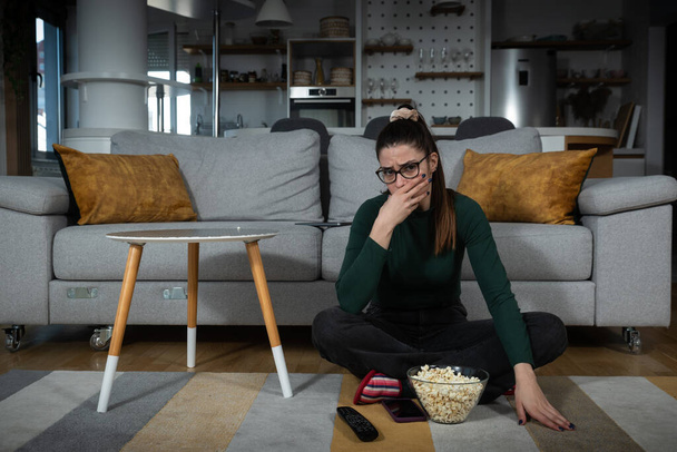 Young scared woman sitting alone at home on the floor watching scary horror movie on the television. Disgusted animal lover female saw animal cruelty tv show feeling sad, disturbed, sorry for animals. - Photo, image