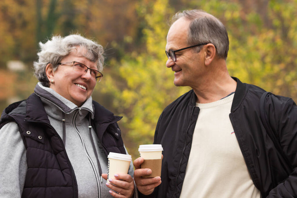 Egy boldog nyugdíjas pár sétál az őszi parkban és kávézik. Mosolygó idős párok beszélgetnek és eldobható poharakat tartanak, kávét elvitelre. Hétvégi szabadidős tevékenység az idősek számára koncepció. Aktív élet. - Fotó, kép