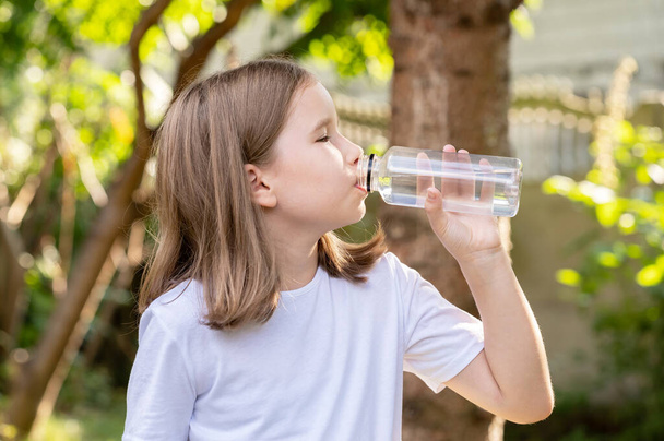 Peruskoulun ikäinen lapsi, tyttö juo tuoretta puhdasta kirkasta suodatettua vettä uudelleenkäytettävästä läpinäkyvästä muovipullosta ulkona muotokuva yksi henkilö Lapsen juomavesi, virvoke, nesteytys käsite - Valokuva, kuva
