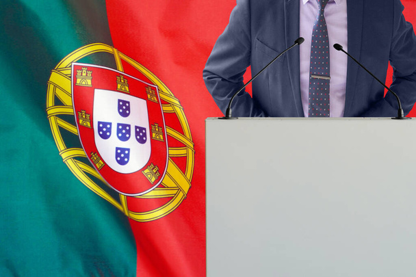 Tribune s mikrofonem a muž v obleku na pozadí portugalské vlajky. Podnikatel a tribun na pozadí portugalské vlajky. Politik na pódiu s mikrofony pozadí Portugalsko vlajky. Konference - Fotografie, Obrázek