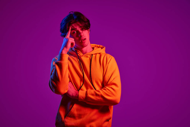 Portrait of young man in casual hoodie posing with thoughtful face isolated over purple background in neon light. Concept of youth, casual fashion, facial expression, emotions, lifestyle, ad - Foto, Imagem