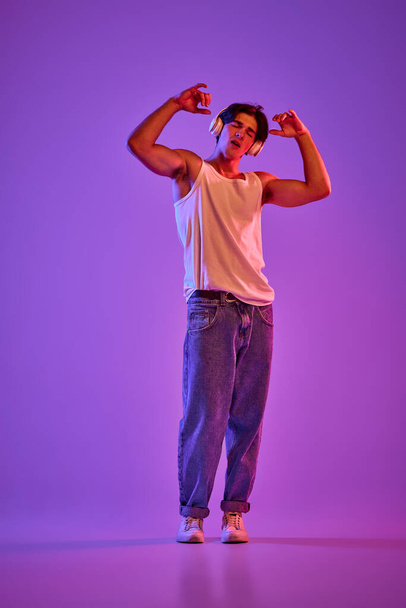 Portrait of young man in white T-shirt, singlet posing, listening to music in headphones over purple background in neon. Concept of youth, casual fashion, facial expression, emotions, lifestyle, ad - Photo, Image