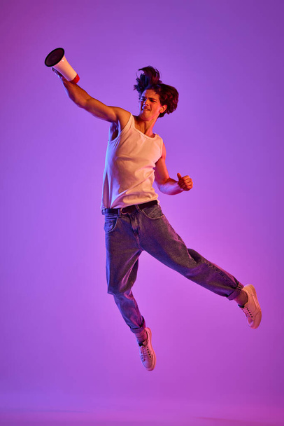 Portrét mladého muže v bílém tričku, jak v neonovém světle pózuje ve skoku přes purpurové pozadí. Megafonové zprávy. Koncept mládí, ležérní móda, výraz obličeje, emoce, životní styl, reklama - Fotografie, Obrázek