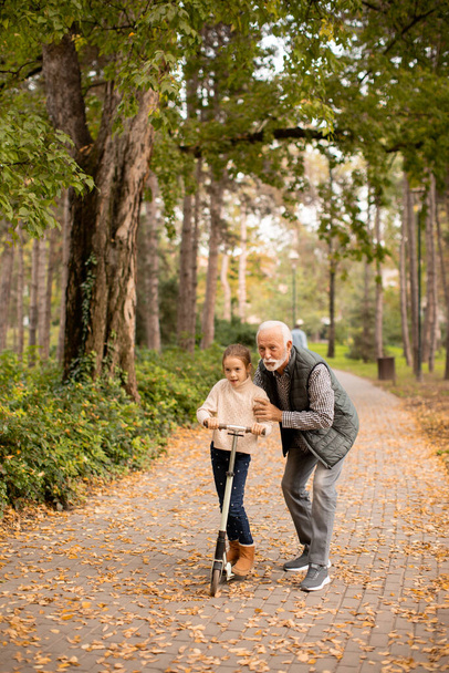 Starší muž učí svou vnučku, jak jezdit na koloběžce v podzimním parku - Fotografie, Obrázek