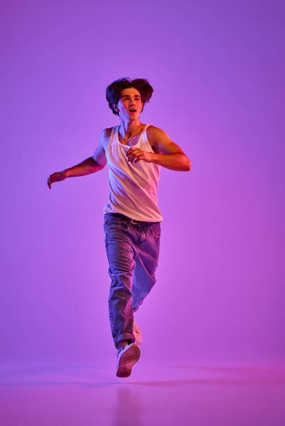 Portrait of young muscular man in white T-shirt, singlet posing in a run over purple background in neon light. Concept of youth, casual fashion, facial expression, emotions, lifestyle, ad - Фото, зображення