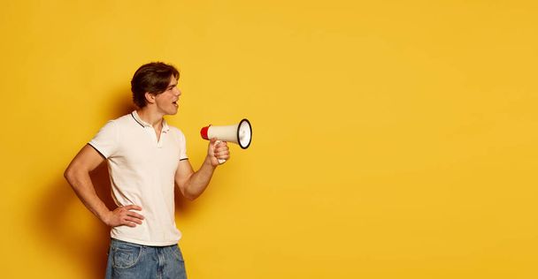 Portrait of young man in casual white T-shirt posing, talking in megaphone over yellow background. Flyer. News, sales . Concept of youth, casual fashion, facial expression, emotions, lifestyle, ad - Photo, Image