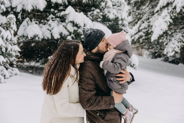 Mutter und Vater halten kleine Tochter im Arm, draußen an einem verschneiten Wintertag, küssen ihre Nase. - Foto, Bild