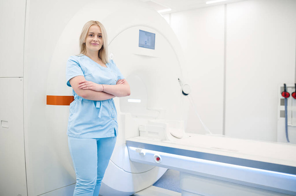 MRI scanner. Blonde medical specialist in blue uniform near MRI scanner - Foto, afbeelding