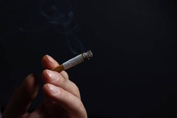 Male hand holding a smoking cigarette on a dark background copy space. harm from smoking cigarettes - Zdjęcie, obraz