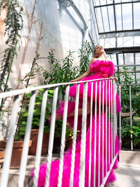 A beautiful young woman with blonde hair in a lush pink tulle dress poses standing on the stairs in a green garden. Style, fashionable girl, beauty. - Photo, Image