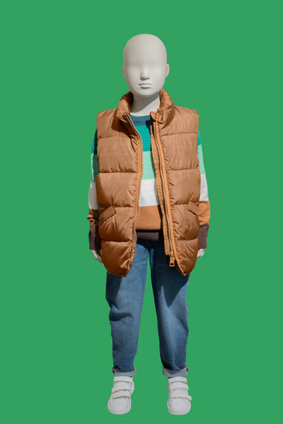 Imagem de comprimento total de um manequim de exibição infantil usando colete acolchoado quente e jeans azuis isolados em um fundo verde - Foto, Imagem