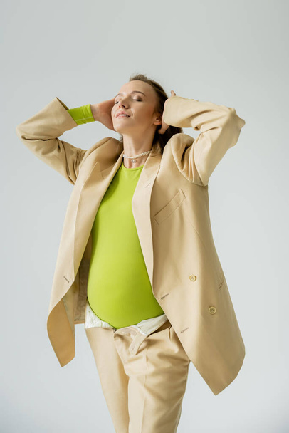 Fashionable pregnant woman in beige jacket touching hair isolated on grey  - Фото, зображення