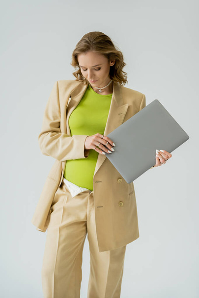 Elegante freelancer embarazada en traje beige que sostiene el ordenador portátil aislado en gris  - Foto, Imagen