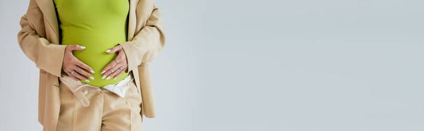 Обрезанный вид беременной женщины в бежевом костюме, касающейся живота, изолированного на сером, баннере  - Фото, изображение