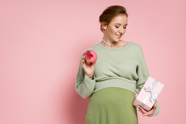 Χαρούμενη έγκυος γυναίκα που κρατά γλυκό ντόνατ και δώρο σε ροζ φόντο  - Φωτογραφία, εικόνα