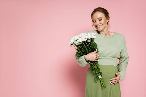 Positive pregnant woman in green outfit looking at bouquet on pink background  - Foto, immagini