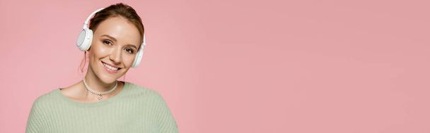 Positive woman in headphones and green sweater looking at camera isolated on pink, banner  - Foto, imagen