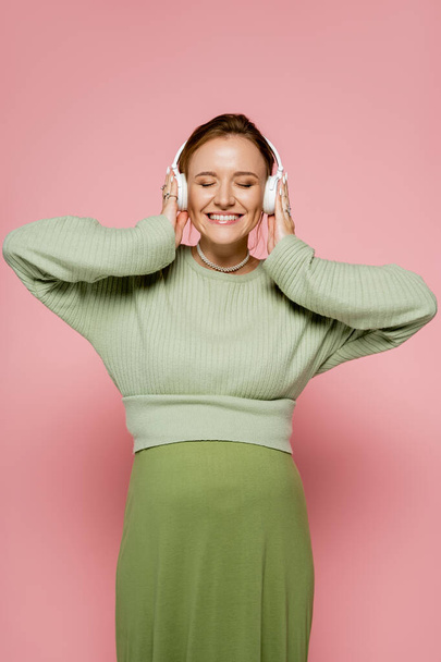 Cheerful pregnant woman in sweater listening music in headphones on pink background  - Фото, изображение