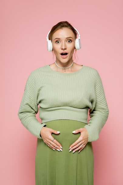 Shocked pregnant woman in green outfit touching belly and using headphones isolated on pink  - Fotoğraf, Görsel