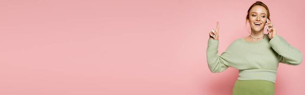 Positive pregnant woman talking on smartphone and pointing with finger on pink background, banner  - Foto, Bild
