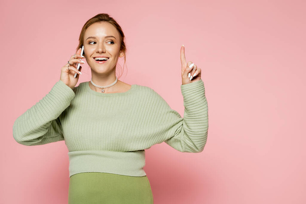 Lächelnde Schwangere im Pullover spricht auf Handy und zeigt mit dem Finger auf rosa Hintergrund  - Foto, Bild