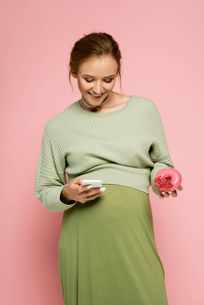Беременная женщина в зеленом костюме с помощью смартфона и держа пончик изолирован на розовый  - Фото, изображение