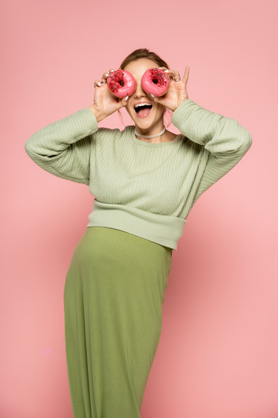 Mujer embarazada emocionada cubriendo la cara con rosquillas sobre fondo rosa  - Foto, imagen