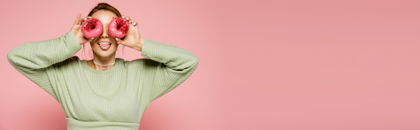 Femme en pull vert sortant la langue et tenant des beignets près du visage isolés sur rose, bannière  - Photo, image